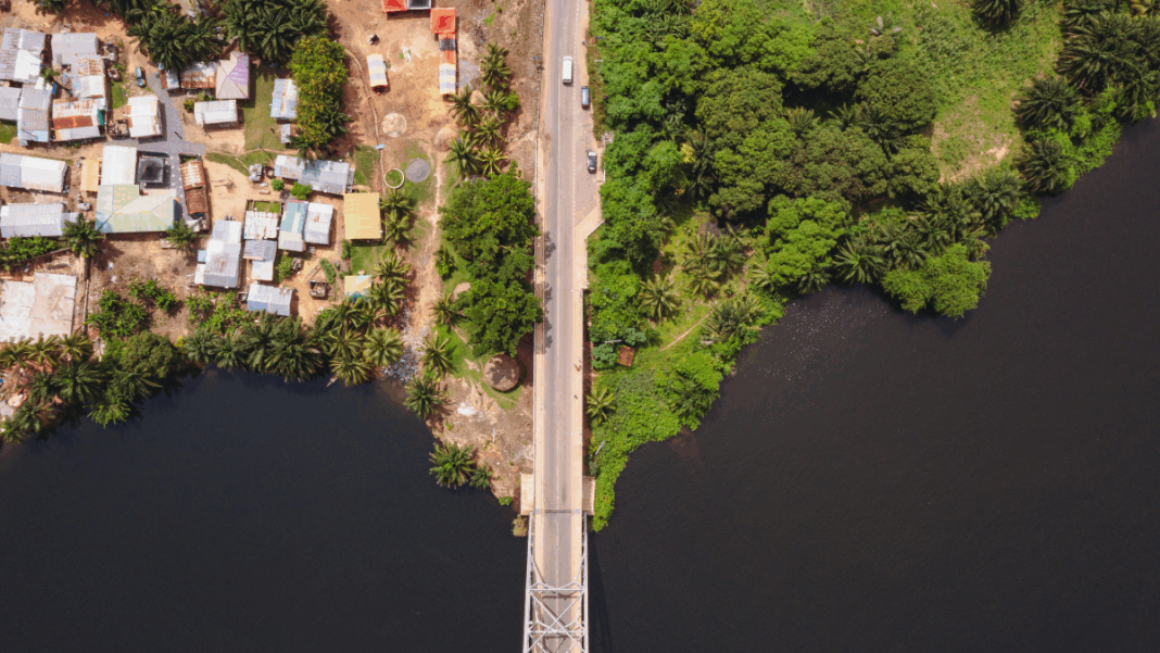 Imagem aérea mostra uma ponte estreita cruzando um rio escuro, ligando duas margens contrastantes — à esquerda, uma área urbana com casas simples e telhados coloridos; à direita, uma região de vegetação densa e verde. A cena evidencia a transição entre ambiente urbano e natureza.