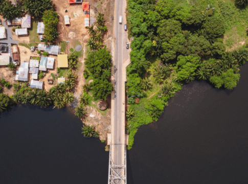 Imagem aérea mostra uma ponte estreita cruzando um rio escuro, ligando duas margens contrastantes — à esquerda, uma área urbana com casas simples e telhados coloridos; à direita, uma região de vegetação densa e verde. A cena evidencia a transição entre ambiente urbano e natureza.