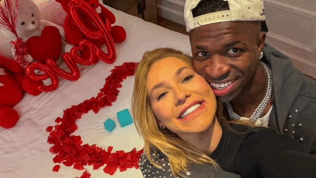 Vinícius Júnior e Virginia estão abraçados à frente de uma cama com pétalas de rosa e balões vermelhos