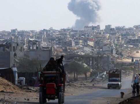 Trator carrega duas pessoas em estrada enquanto ao fundo uma fumaça grande aparece entre as casas