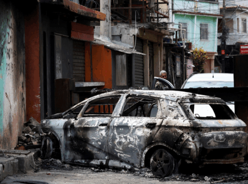 Homem está atrás de carro destruído por fogo na entrada da Favela da Penha
