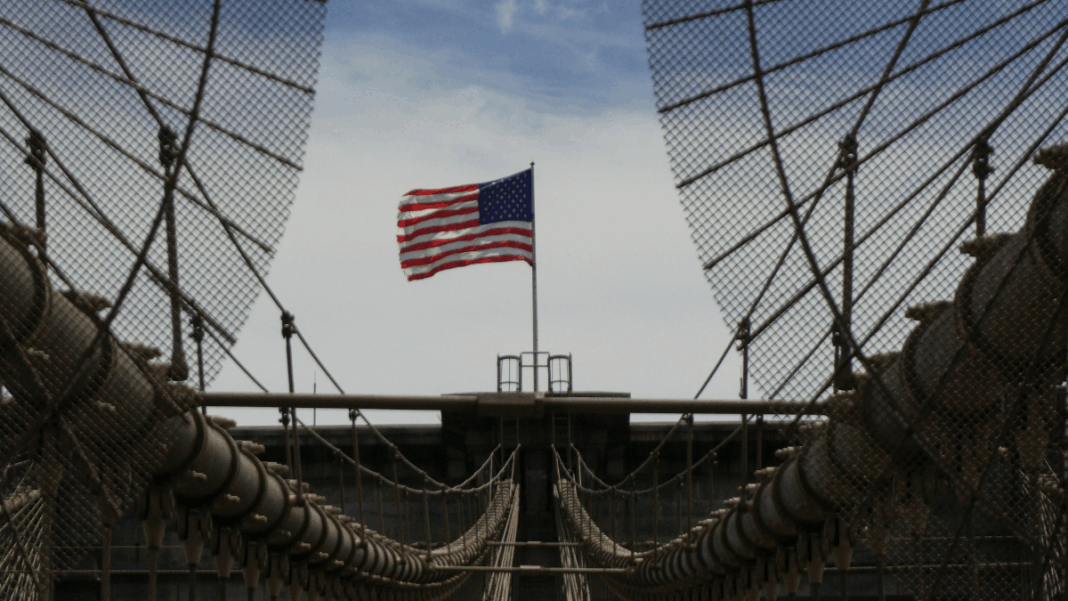 Fotografia tirada de uma ponte suspensa com cabos de aço e grades metálicas convergindo para o centro da imagem. Ao fundo, no ponto mais alto da estrutura, tremula uma bandeira dos Estados Unidos sob um céu parcialmente nublado. A composição destaca as linhas simétricas da ponte e o contraste entre o aço e o movimento suave da bandeira.