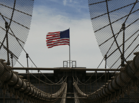 Fotografia tirada de uma ponte suspensa com cabos de aço e grades metálicas convergindo para o centro da imagem. Ao fundo, no ponto mais alto da estrutura, tremula uma bandeira dos Estados Unidos sob um céu parcialmente nublado. A composição destaca as linhas simétricas da ponte e o contraste entre o aço e o movimento suave da bandeira.