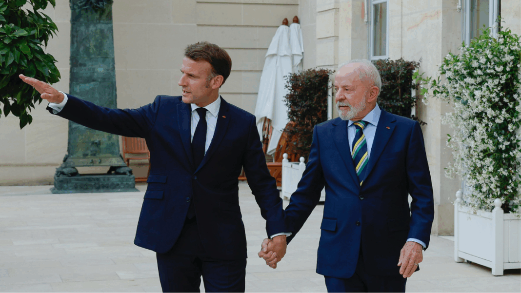 Dois homens de terno escuro estão de mãos dadas e caminhando em uma área externa elegante, com plantas e paredes de pedra clara ao fundo. O homem à esquerda gesticula com o braço estendido, apontando algo à frente, enquanto o homem à direita olha na mesma direção com expressão atenta. Ambos parecem estar em um ambiente formal e amistoso.
