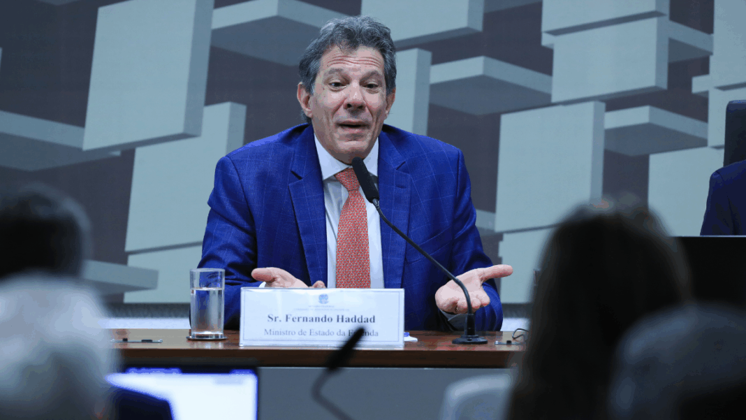 Homem de terno azul e gravata vermelha fala ao microfone em uma mesa de audiência. À sua frente há uma placa com o nome “Sr. Fernando Haddad, Ministro de Estado da Fazenda”. Ele gesticula com as mãos abertas, em gesto de explicação. Ao fundo, há um painel com formas geométricas e pessoas assistindo à fala.