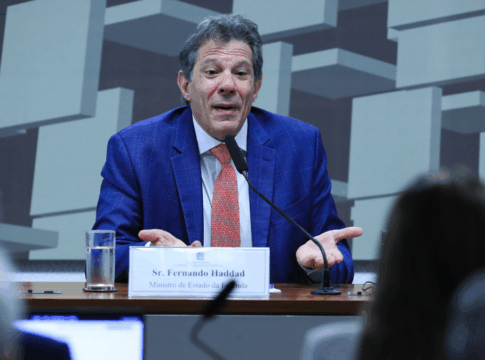 Homem de terno azul e gravata vermelha fala ao microfone em uma mesa de audiência. À sua frente há uma placa com o nome “Sr. Fernando Haddad, Ministro de Estado da Fazenda”. Ele gesticula com as mãos abertas, em gesto de explicação. Ao fundo, há um painel com formas geométricas e pessoas assistindo à fala.