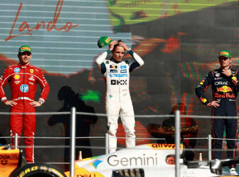 Três pilotos de Fórmula 1 estão no pódio após uma corrida. No centro, o vencedor, vestido com macacão branco da equipe McLaren, segura o boné verde da Pirelli sobre a cabeça. À esquerda, um piloto da Ferrari, de macacão vermelho, observa com as mãos na cintura. À direita, um piloto da Red Bull, de macacão azul escuro, mantém a mão no queixo. Em primeiro plano, parte do carro laranja e preto da McLaren aparece, e ao fundo, um telão exibe o nome “Lando”, indicando o vencedor da prova.