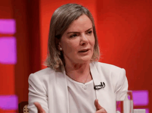 Mulher de cabelos loiros curtos veste blazer branco e fala durante uma entrevista ou debate em estúdio com fundo vermelho e luzes roxas. Ela gesticula com as mãos e usa microfone preso à roupa.