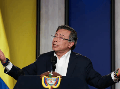 Homem de terno escuro e camisa branca fala em um púlpito com o brasão da Colômbia. Ele está com os braços abertos e expressão séria, olhando para o alto. Usa óculos e várias pulseiras coloridas nos pulsos. Ao fundo, há um painel com as cores da bandeira colombiana — amarelo, azul e vermelho —, sugerindo um pronunciamento oficial.