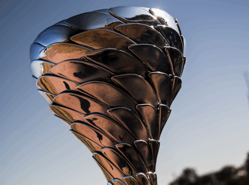 Troféu metálico prateado com design moderno, formado por placas sobrepostas que lembram escamas ou pétalas. Ele está exposto sobre uma base preta, com um autódromo ao fundo sob céu azul e luz solar intensa, sugerindo um evento de corrida ou premiação esportiva.