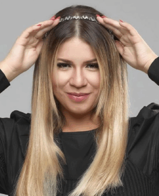 Mulher de cabelos lisos e loiros posa sorrindo levemente diante de um fundo cinza. Ela usa uma blusa preta de mangas bufantes e segura uma tiara prateada sobre a cabeça com as duas mãos.