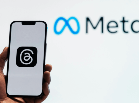 Uma pessoa segura um smartphone que exibe o logotipo do aplicativo Threads, da Meta, em fundo branco. Ao fundo, desfocado, aparece o logotipo da empresa Meta, com o símbolo azul em forma de infinito ao lado da palavra “Meta” em letras pretas. A imagem sugere o vínculo entre o aplicativo e a empresa controladora das redes sociais Facebook e Instagram.