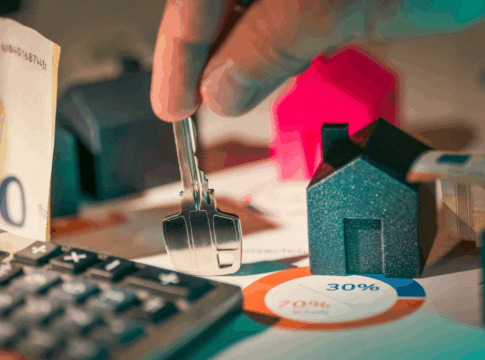 Pessoa segura uma chave sobre uma mesa com miniaturas de casas, notas de dinheiro, calculadora e gráfico circular, representando o mercado imobiliário e o custo do aluguel.