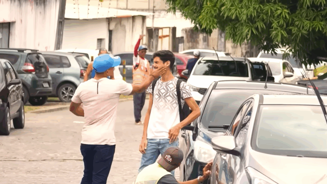 Em uma rua com carros estacionados, um homem de boné azul e camiseta clara dá um tapa no rosto de outro homem mais jovem, que usa mochila e camiseta branca com estampa geométrica. O jovem parece surpreso e inclina o rosto para o lado. Ao fundo, há outras pessoas caminhando e um homem com colete laranja acenando com o braço levantado. A cena ocorre em ambiente urbano, durante o dia
