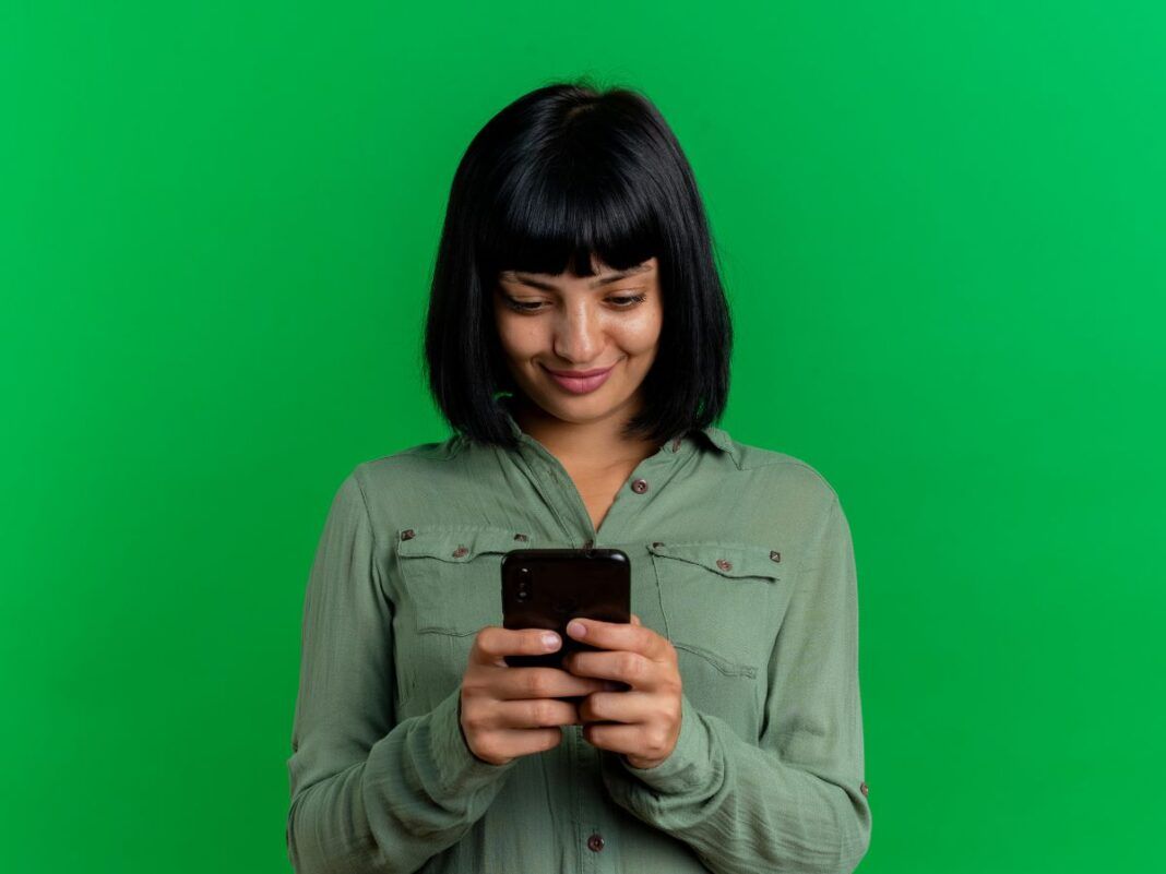 Mulher de cabelo preto com franja em um fundo verde segurando um celular