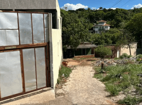 Entrada de uma casa simples com portão metálico em uma rua de terra, cercada por vegetação e outras residências ao fundo, em área residencial de periferia.