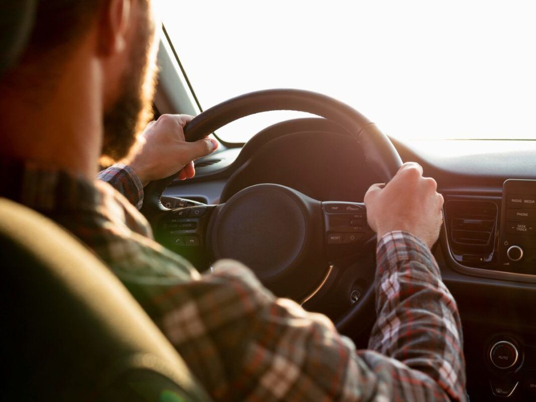 A imagem mostra um homem de costas dirigindo com uma camisa xadrez e as duas mãos no volante
