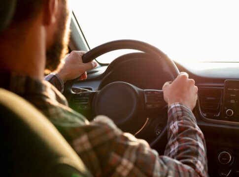 A imagem mostra um homem de costas dirigindo com uma camisa xadrez e as duas mãos no volante
