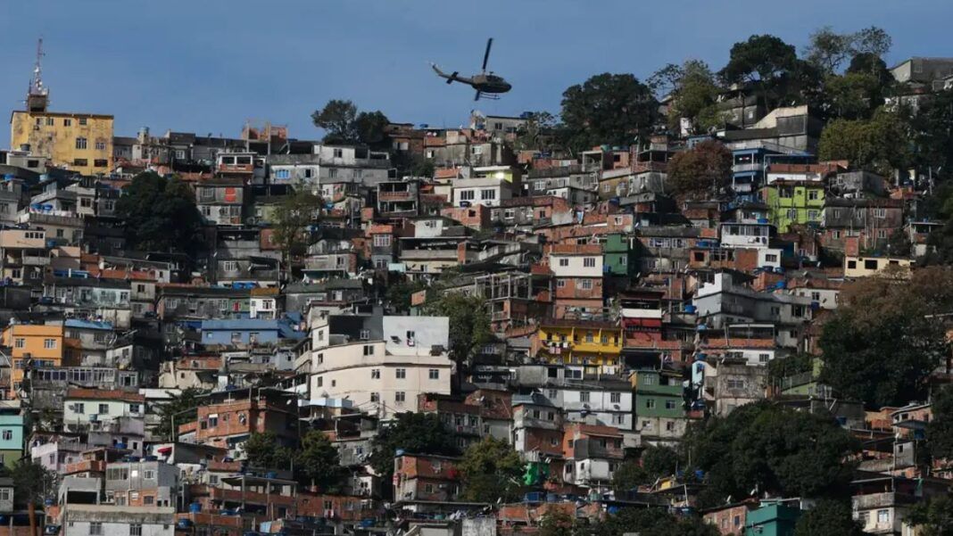 Uma foto panorâmica, tirada em plano aberto, que mostra uma grande favela ou comunidade verticalizada no Rio de Janeiro, estendendo-se por um morro. A cena é capturada em um dia claro, com céu azul.
