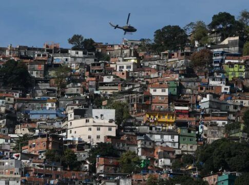 Uma foto panorâmica, tirada em plano aberto, que mostra uma grande favela ou comunidade verticalizada no Rio de Janeiro, estendendo-se por um morro. A cena é capturada em um dia claro, com céu azul.