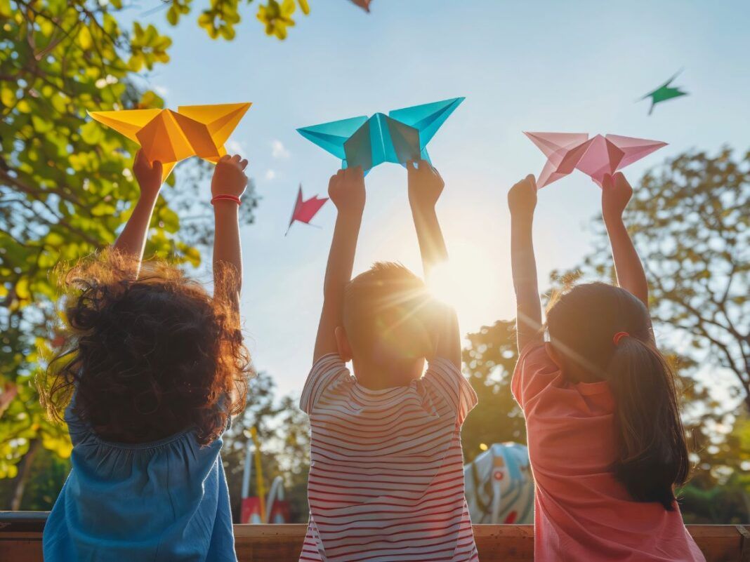 A imagem mostra três crianças brincando de costas em um parque com origamis nas mãos