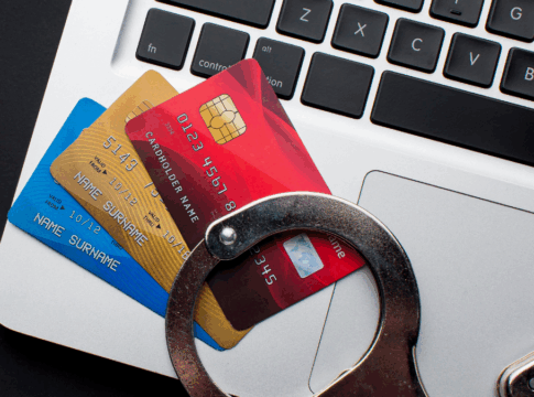 Cartões de crédito coloridos sobre o teclado de um notebook prateado, com um par de algemas apoiado por cima, simbolizando crimes financeiros e fraudes bancárias online