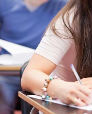 Aluna sentada em uma carteira de escola com uma caneta na mão, escrevendo em uma folha