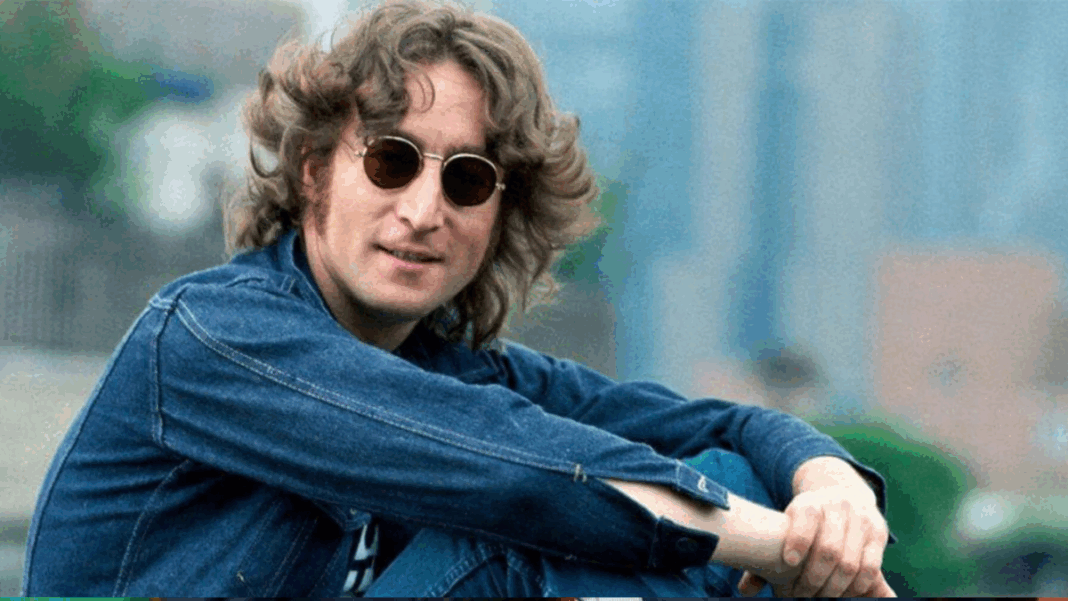 Johnn Lennon sentado de lado, em um espaço aberto e gramado, com camisa jeans e óculos de sol redondo.
