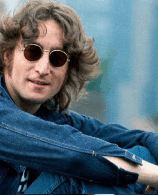 Johnn Lennon sentado de lado, em um espaço aberto e gramado, com camisa jeans e óculos de sol redondo.