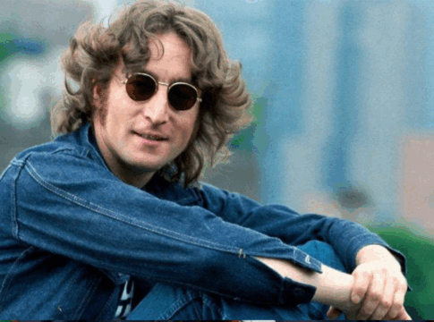 Johnn Lennon sentado de lado, em um espaço aberto e gramado, com camisa jeans e óculos de sol redondo.