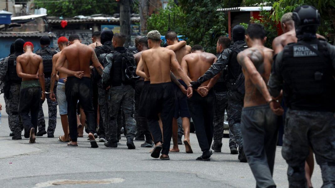 A fotografia, capturada em um ambiente de rua, numa comunidade, mostra um grupo de homens, alguns deles detidos, sendo conduzidos por policiais militares.