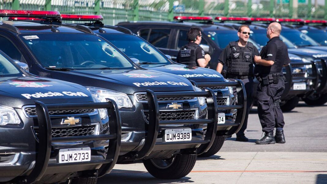 Viaturas estão enfileiradas num estacionamento. Ao lado dos veículos policiais, aparecem três policiais civis.