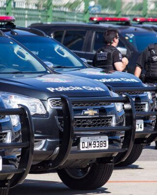 Viaturas estão enfileiradas num estacionamento. Ao lado dos veículos policiais, aparecem três policiais civis.