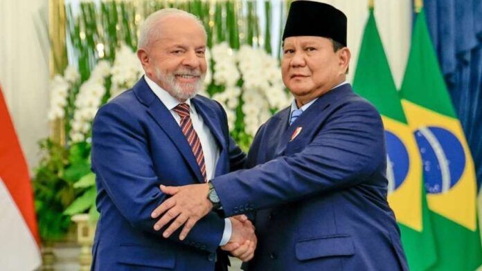 Uma foto em plano médio e em ambiente interno registra o aperto de mãos entre o Presidente do Brasil, Luiz Inácio Lula da Silva (à esquerda), e o Presidente da Indonésia, Prabowo Subianto (à direita), durante um encontro oficial, em Jacarta. No fundo, notam-se as bandeiras nacionais, ligeiramente desfocadas: a bandeira da Indonésia (vermelha e branca) e a bandeira do Brasil (verde, amarela e azul), confirmando o contexto diplomático da reunião. A iluminação é forte, de ambiente formal, com decorações florais brancas visíveis atrás dos líderes.