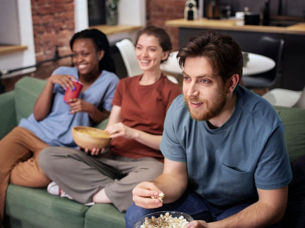 Um homem e duas mulheres estão sentados em um sofá com pipoca na mão e olhando para uma TV