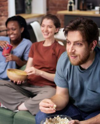 Um homem e duas mulheres estão sentados em um sofá com pipoca na mão e olhando para uma TV