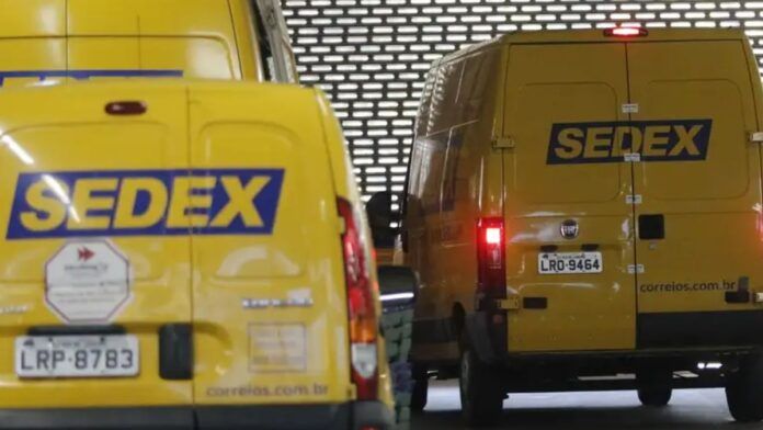 Imagem mostra a traseira de duas vans de entrega dos Correios, ambas na cor amarela característica, em uma área de depósito ou doca de carregamento com uma parede de metal perfurada ao fundo.