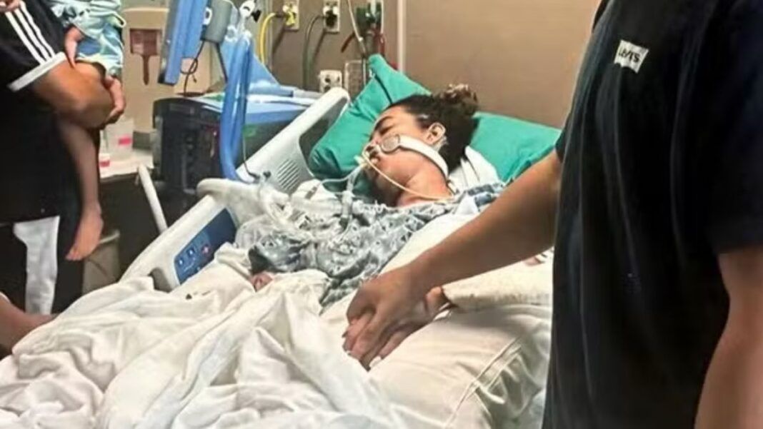 A imagem retrata uma mulher em estado grave na cama de um hospital, com o suporte de equipamentos médicos, enquanto familiares ou a acompanham, segurando na mão dela. A cama é branca com lençóis brancos, e a paciente está coberta por um cobertor estampado em tons de cinza. Vários fios, tubos e monitores estão visíveis ao redor da paciente e da cama, característicos de uma unidade de terapia intensiva ou cuidados críticos.