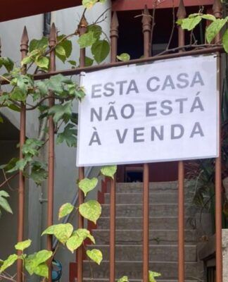 Uma foto de um portão de ferro, coberto por algumas trepadeiras de folhas verdes, que revela a entrada de uma casa. O portão está pintado de marrom, e a parede, ao seu lado direito, é de um tom de vermelho terracota. Pendurado no portão, no centro da imagem, há um cartaz retangular branco com letras pretas em caixa alta, que diz: 