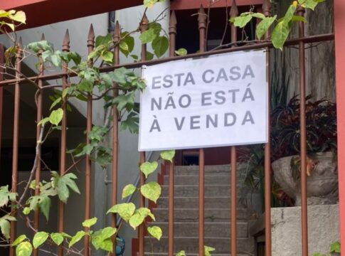 Uma foto de um portão de ferro, coberto por algumas trepadeiras de folhas verdes, que revela a entrada de uma casa. O portão está pintado de marrom, e a parede, ao seu lado direito, é de um tom de vermelho terracota. Pendurado no portão, no centro da imagem, há um cartaz retangular branco com letras pretas em caixa alta, que diz: 