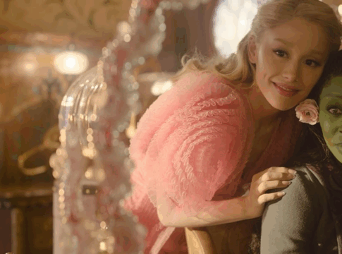 Glinda com a mão no ombro de Elphaba, enquanto ambas se olham no espelho