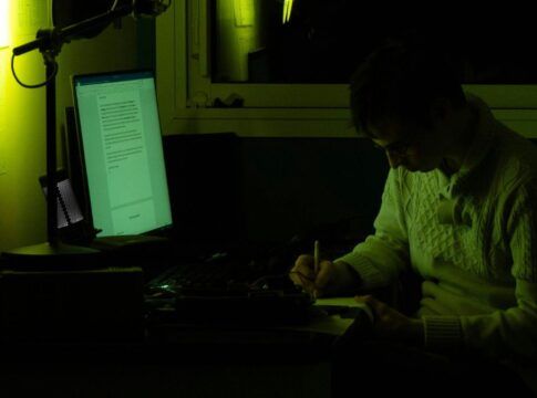 Homem no escuro, com a tela do computador ligada, escrevendo em um caderno à mão