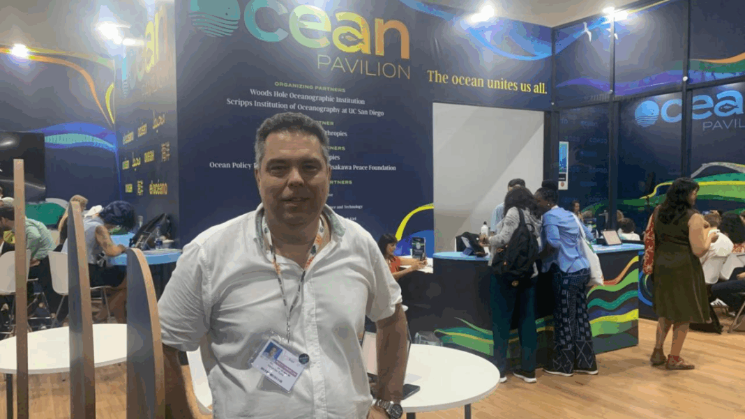Homem usando camisa branca e crachá posa em pé diante do estande “Ocean Pavilion”, onde várias pessoas trabalham e conversam ao fundo; o espaço tem paredes decoradas com tons de azul e verde e a frase “The ocean unites us all.”