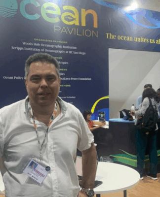 Homem usando camisa branca e crachá posa em pé diante do estande “Ocean Pavilion”, onde várias pessoas trabalham e conversam ao fundo; o espaço tem paredes decoradas com tons de azul e verde e a frase “The ocean unites us all.”