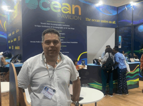 Homem usando camisa branca e crachá posa em pé diante do estande “Ocean Pavilion”, onde várias pessoas trabalham e conversam ao fundo; o espaço tem paredes decoradas com tons de azul e verde e a frase “The ocean unites us all.”