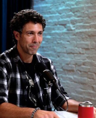 Rodrigo Oliveira sentado em uma mesa, ao lado de uma parede de tijolos brancos