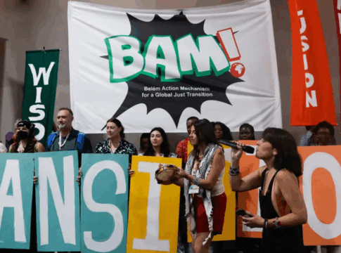Grupo de ativistas segura letras coloridas formando a palavra “TRANSITION” em frente a um grande banner com a inscrição “BAM! Belém Action Mechanism for a Global Just Transition”, enquanto faixas verticais trazem palavras como “Care”, “Wisdom”, “Inclusion” e “Labour Rights” em um ato dentro de espaço fechado.