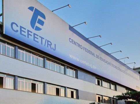 Fachada do prédio do CEFET/RJ, com placas institucionais visíveis, janelas amplas e bandeiras hasteadas, cercado por palmeiras e carros estacionados em frente.