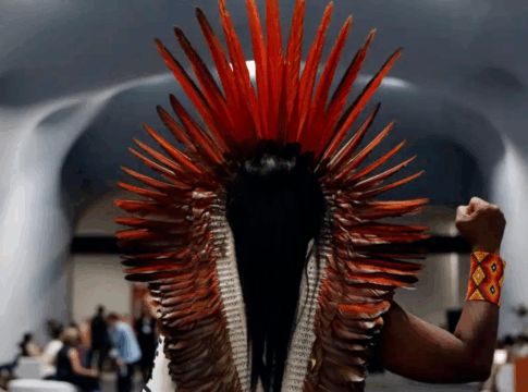 Pessoa indígena vista de costas, usando grande cocar de penas vermelhas e pulseira colorida, ergue o punho direito em gesto de resistência dentro de um salão moderno onde outras pessoas conversam ao fundo.