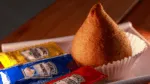 Coxinha dourada colocada em forminha de papel branco sobre bandeja, acompanhada de sachês de maionese, mostarda e ketchup da marca Heinz.